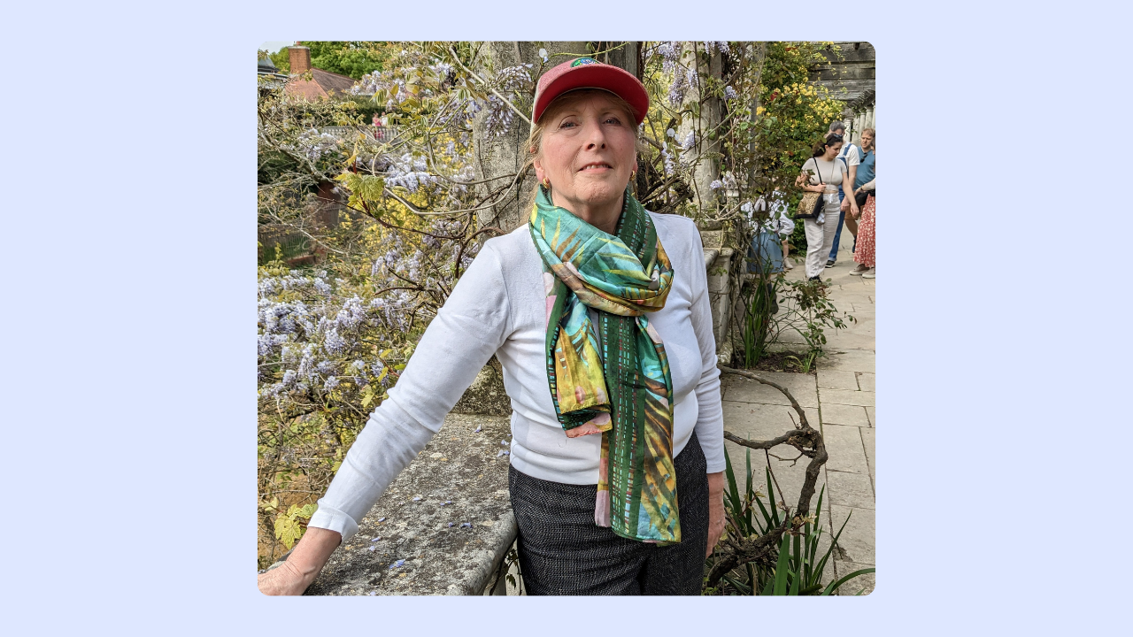 An older woman with a colourful cap and scarf leans on a wall and smiles at the camera, with trees and flowers behind her.