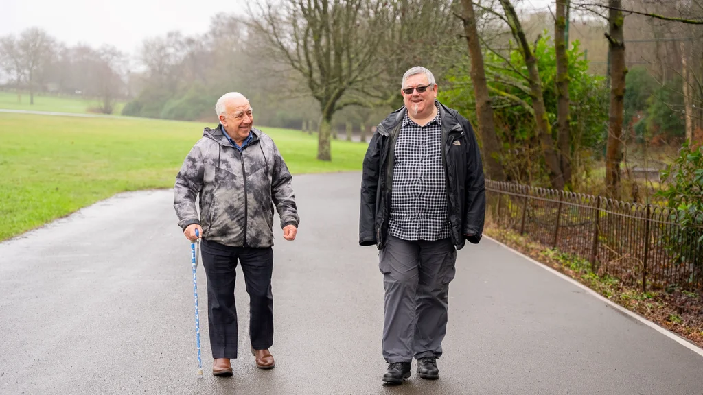 Two older men walking together along a path surrounded by grass and trees.