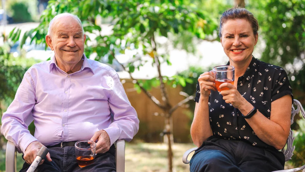 Ann and her father sitting outside holding cups of tea with sunny trees behind them.