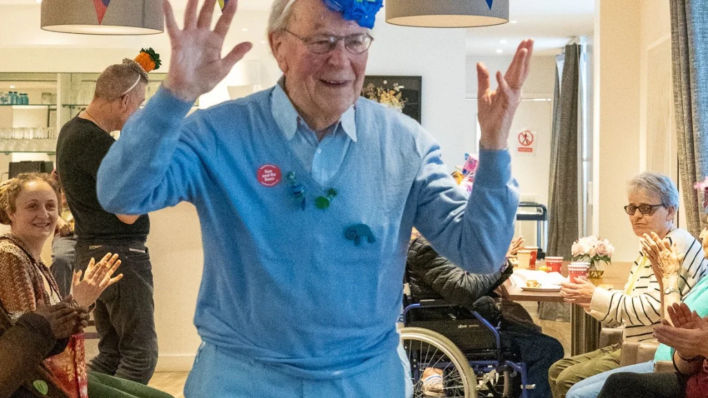 An older man, dressed all in blue with a blue hat, smiles and holds his hands up. In the background there are people sitting and clapping and Age Without Limits bunting is overhead.