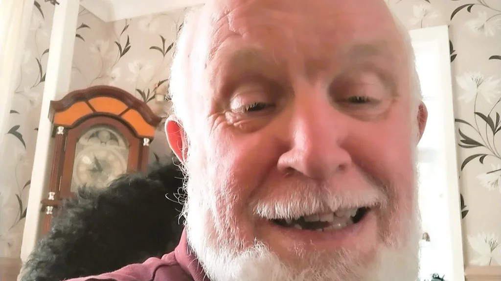 A man with a beard smiles at the camera. Floral patterned wallpaper is in the background.