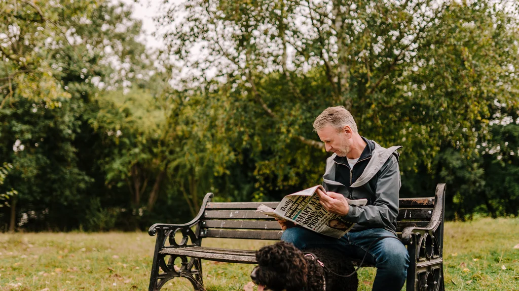 A man with grey hair sits on a bench with a dog and reads a newspaper.