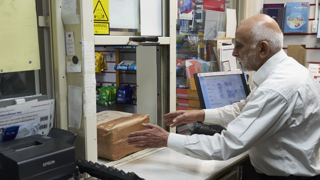 A man sits by a computer and handles a parcel behind the desk at a post office.