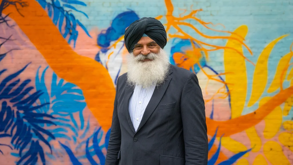 A man with a grey beard and black turban wears a suit and smiles at the camera, standing in front of a wall of art.