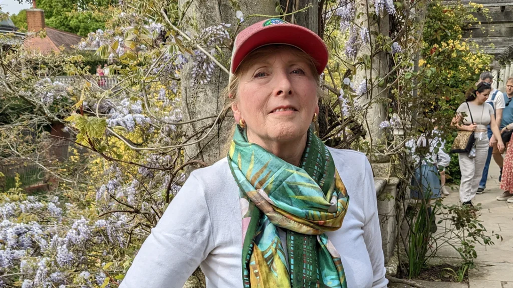 An older woman with a colourful cap and scarf leans on a wall and smiles at the camera, with trees and flowers behind her.